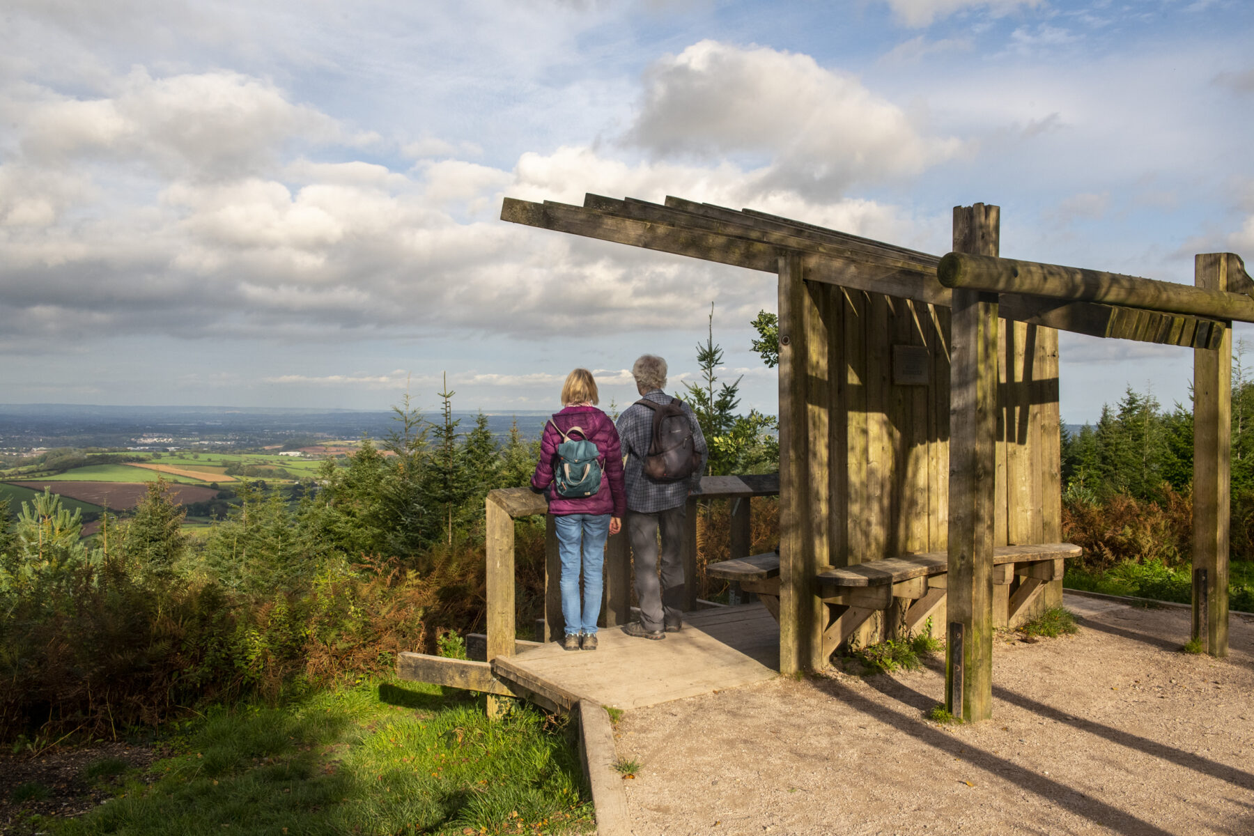 A couple stand under a wooden shelter looking at a panoramic view