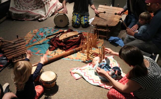 a mother with her baby on the floor holding an instrument, and another toddler to the left playing African drums