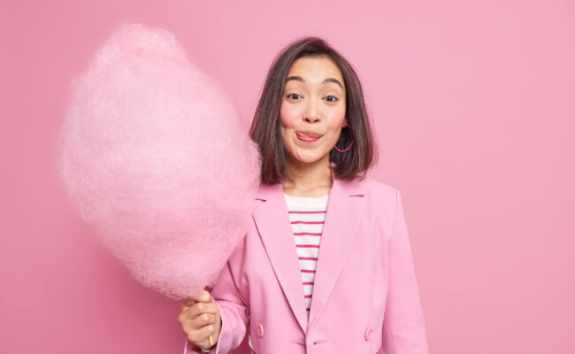 a young woman holding pink candyfloss sticking out her tongue. she is wearing a pink blazer and standing against a pink background